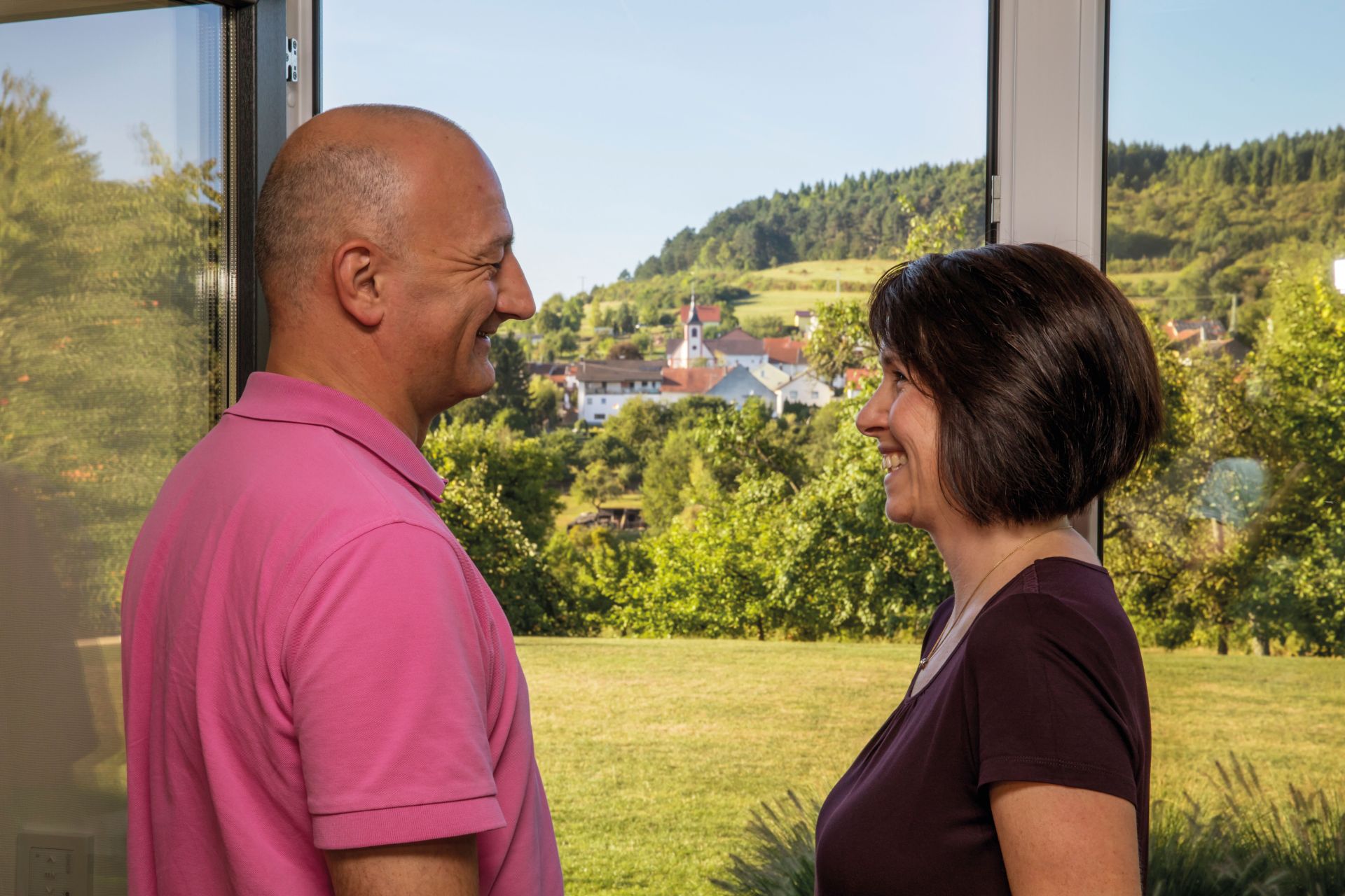 Ein Paar steht sich in einem hellen Raum gegenüber und lächelt sich an, im Hintergrund ist durch das Fenster eine grüne Hügellandschaft zu sehen.