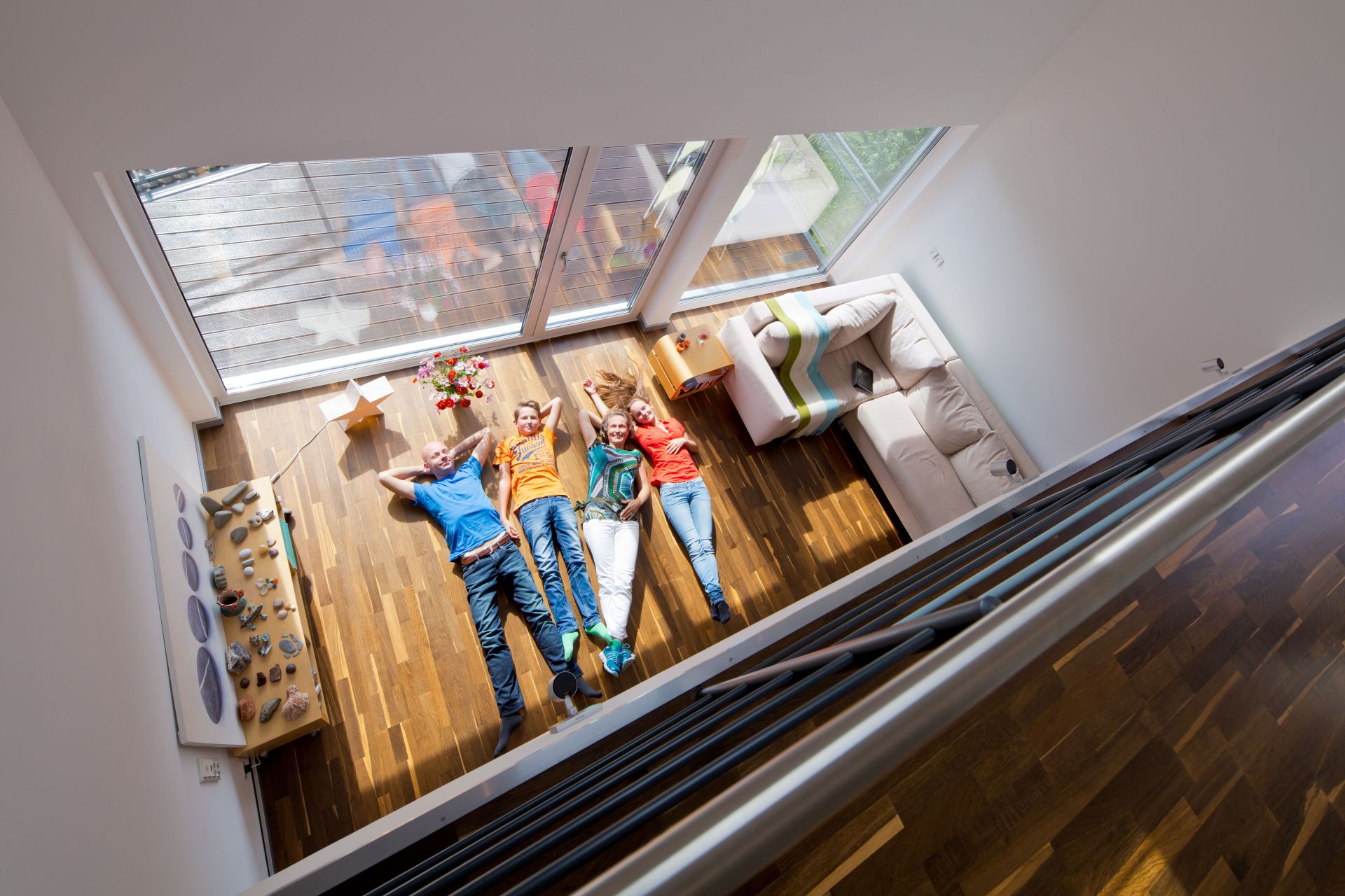 Familie liegt entspannt auf dem Holzboden des hellen Wohnzimmers mit Galerie und großen Panoramafenstern.