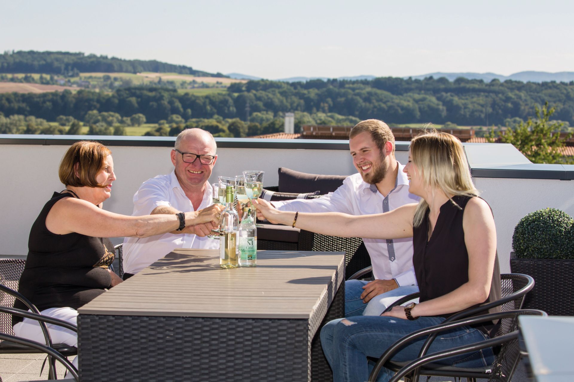 Vier Personen stoßen auf der sonnigen Dachterrasse ihres FingerHaus mit Getränken an und genießen den Ausblick ins Grüne.