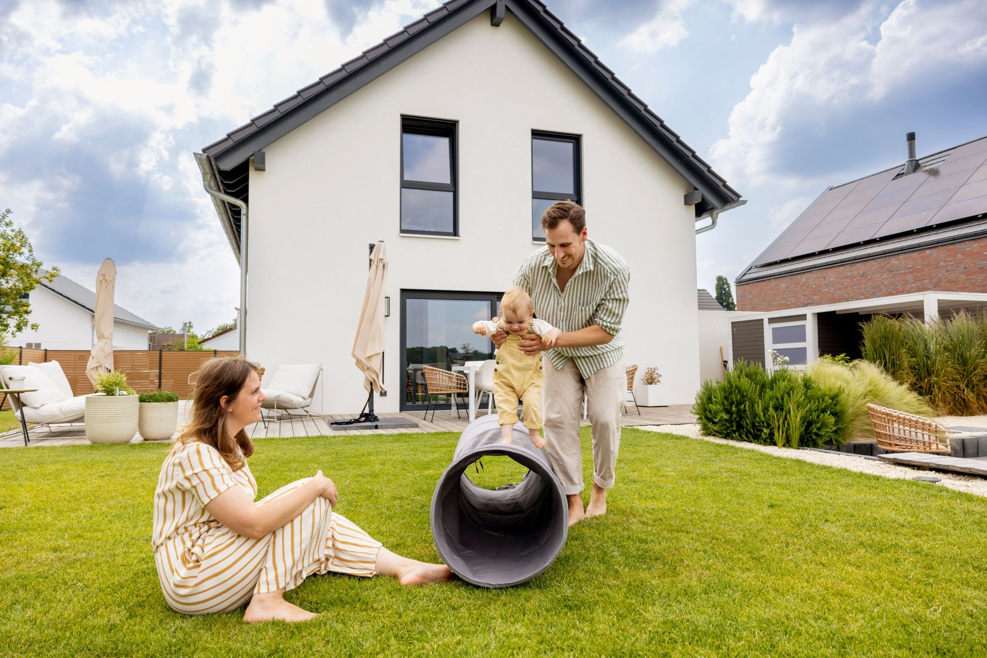 Eine junge Familie spielt barfuß im Garten ihres modernen FingerHaus-Hauses. Vater hilft dem Baby beim Krabbeln über ein Spieltunnel, während die Mutter lachend zusieht.