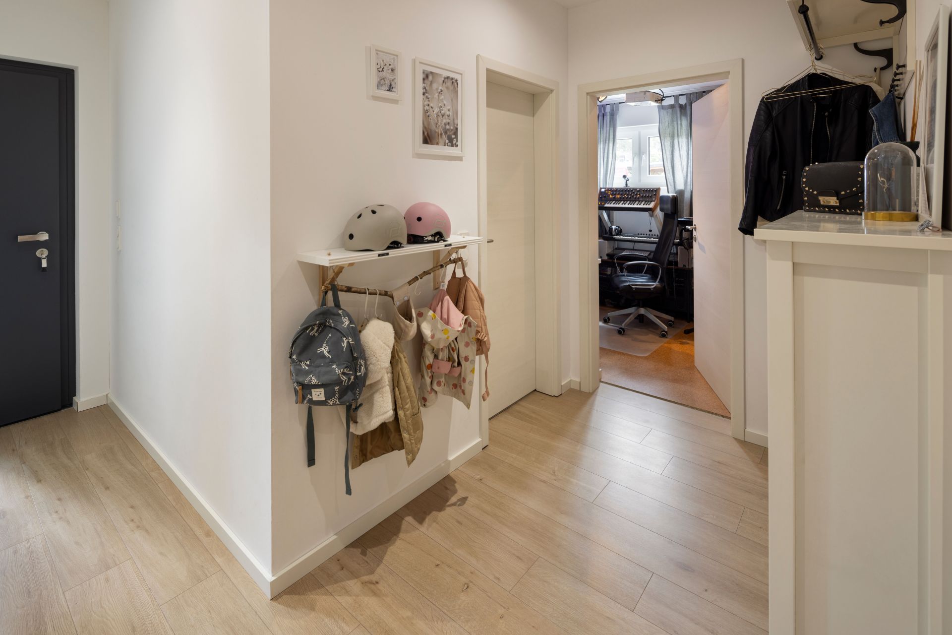 Heller Flur in einem frei geplanten FingerHaus mit Holzfußboden, Garderobe für Kinder und Blick in ein modernes Arbeitszimmer mit Musikequipment.