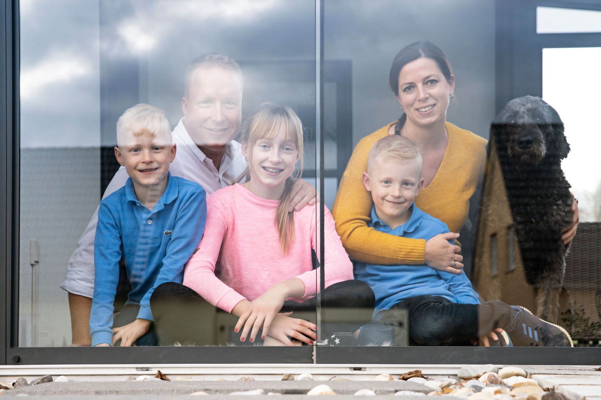 Familie mit zwei Kindern und Hund sitzt gemütlich hinter einer großen Glasfront und blickt nach draußen.