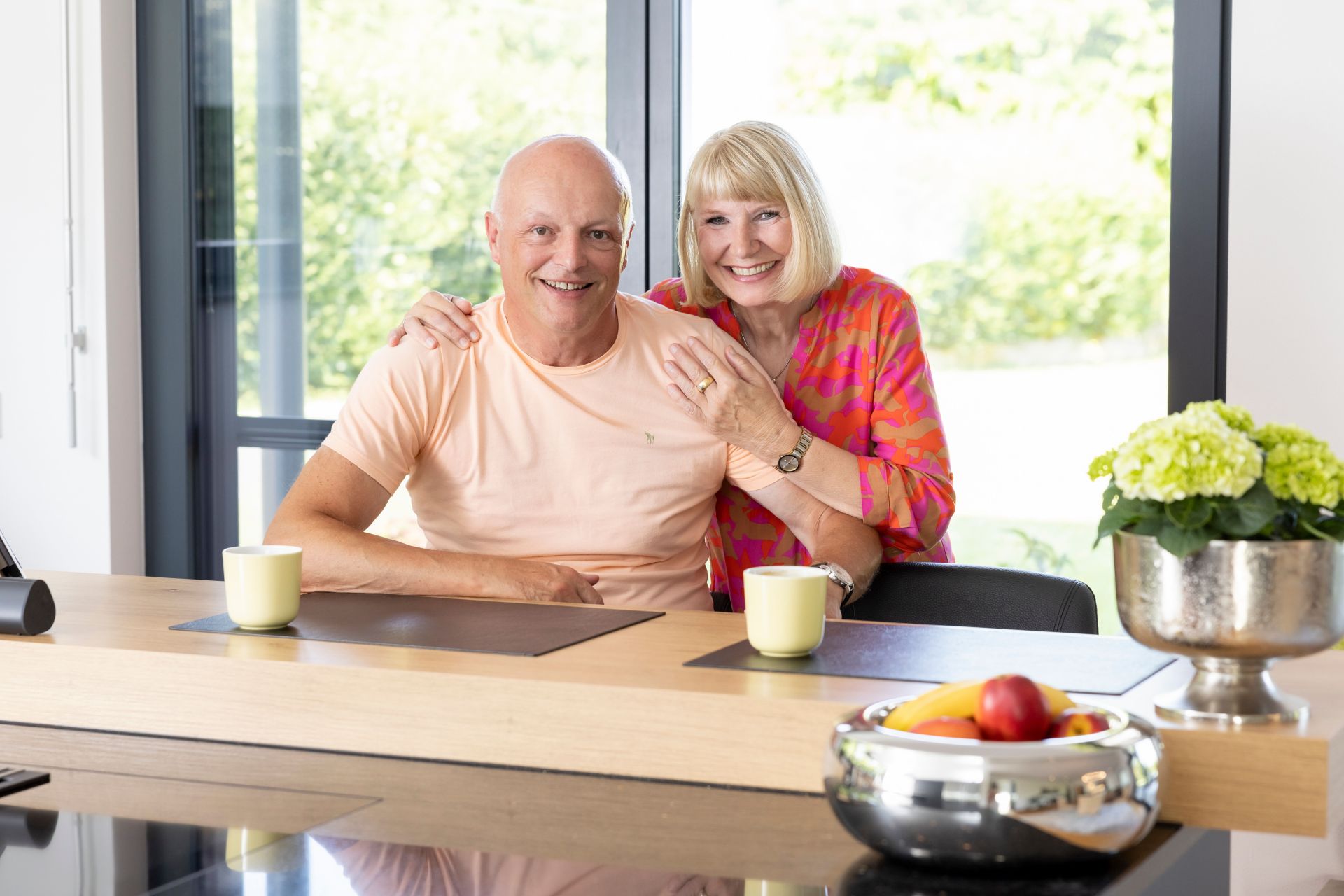 Ein Paar sitzt lächelnd am Küchentresen in einem frei geplanten FingerHaus. Auf dem Tisch stehen Kaffeetassen, eine Obstschale und eine Blumendekoration.
