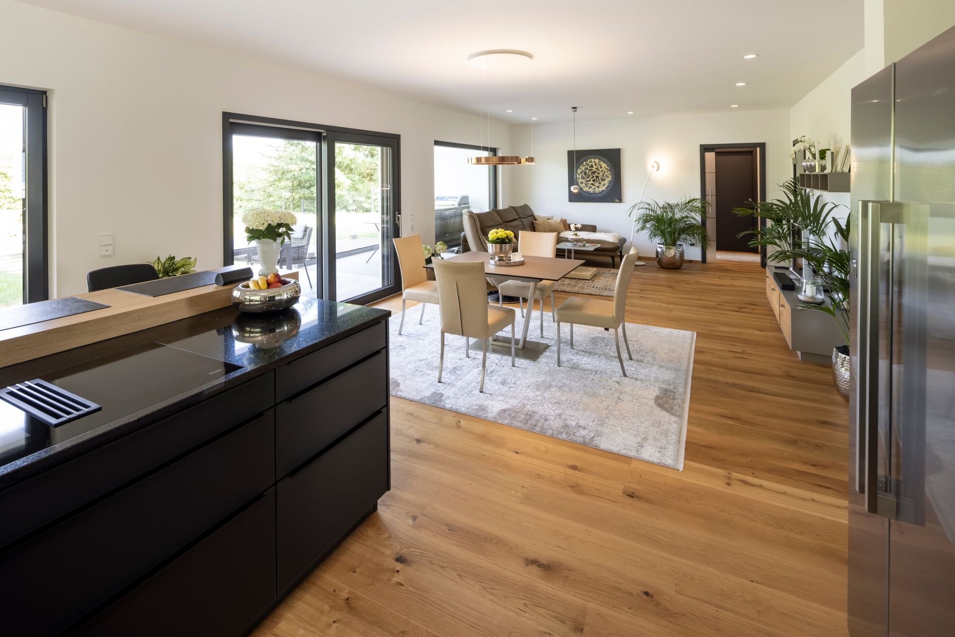 Offener Wohn-, Ess- und Kochbereich in einem frei geplanten FingerHaus. Moderne schwarze Küche mit Kochinsel, angrenzender Essbereich mit großem Tisch und helles Wohnzimmer mit Sofa. Heller Holzboden und große Fenster sorgen für ein freundliches Ambiente.