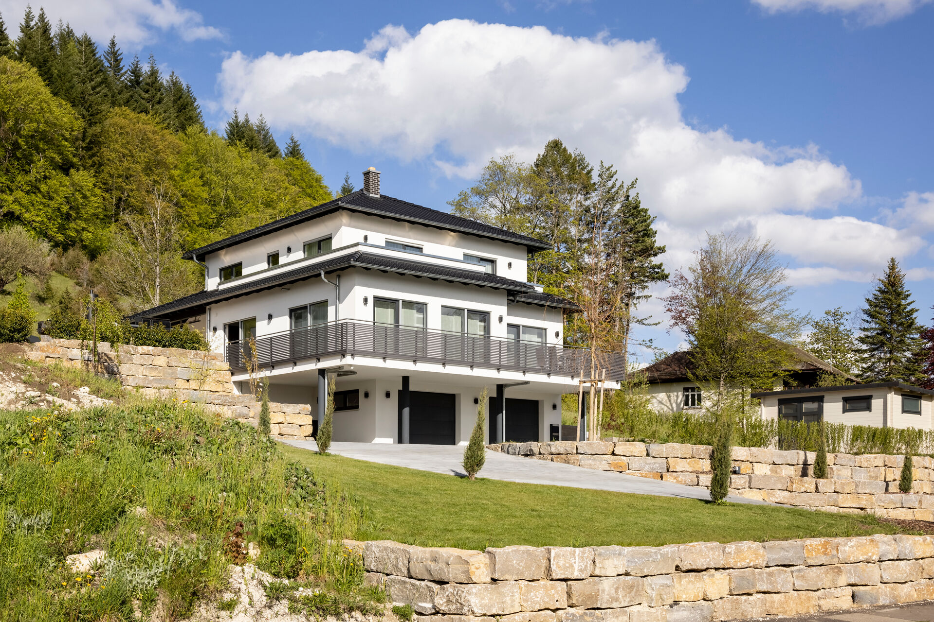 Weißes Einfamilienhaus in Hanglage mit dunklem Walmdach, umlaufendem Balkon, Garage und terrassiertem Garten mit Natursteinmauern.