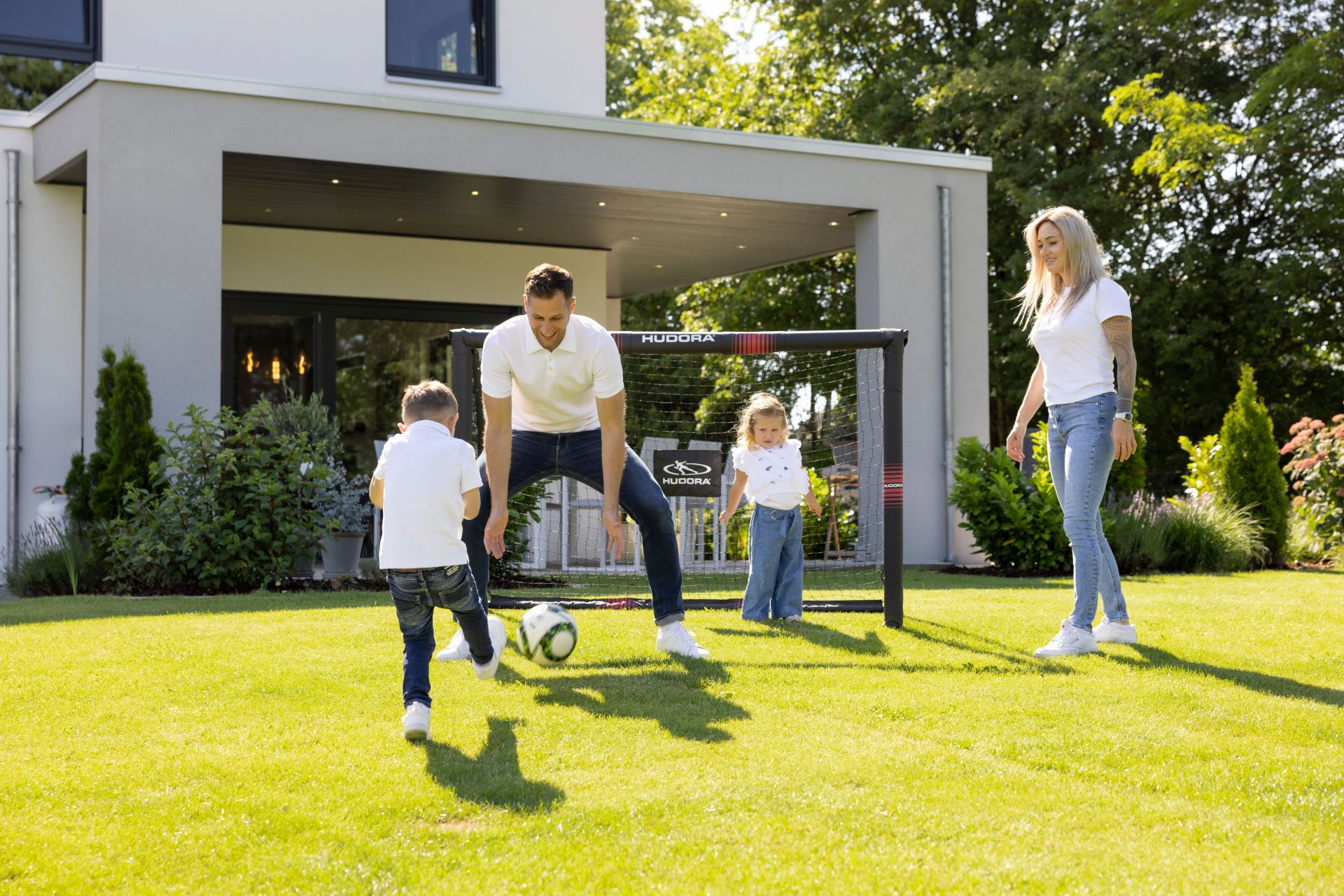 Eine Familie spielt gemeinsam Fußball im Garten eines modernen FingerHaus-Hauses. Vater und Sohn kicken den Ball, während Mutter und Tochter lachend zusehen.