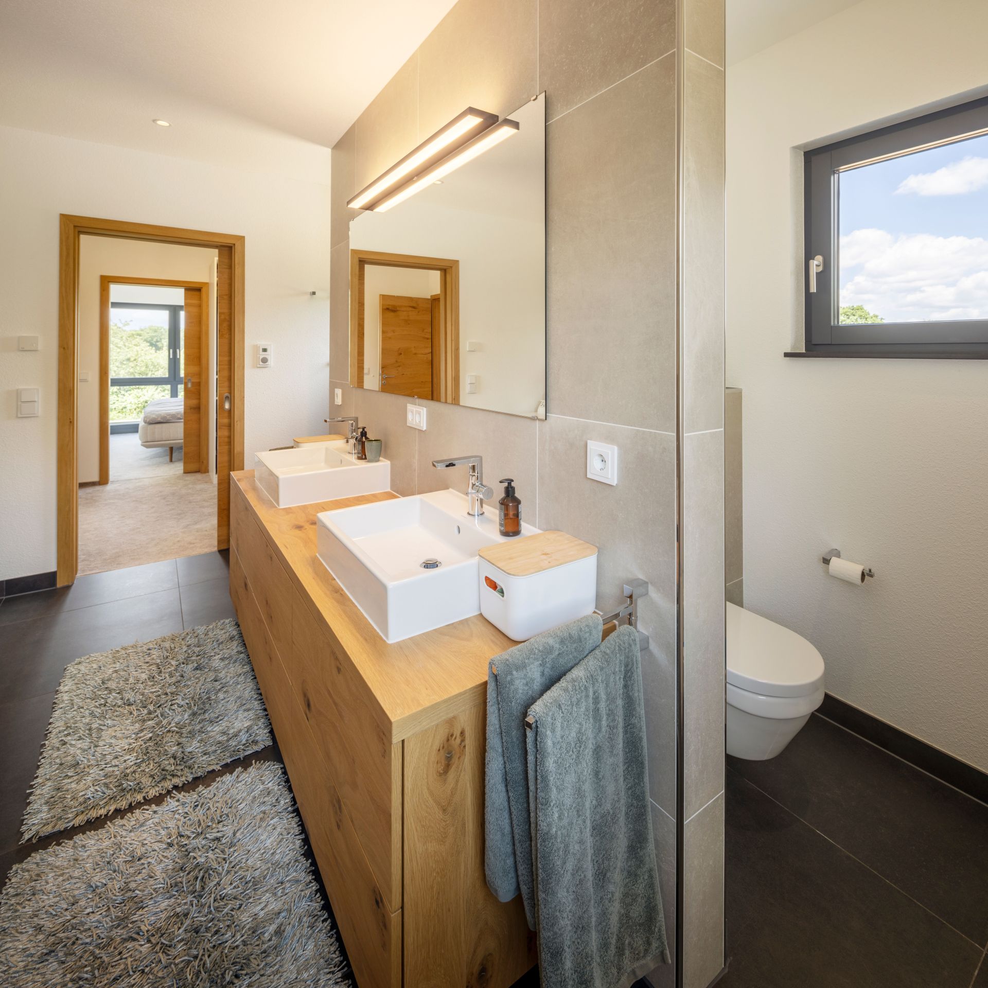 Modernes Badezimmer in einem FingerHaus-Haus mit Doppelwaschbecken auf einer Holzkonsole, großem Spiegel, hellen Fliesen und Fenster mit Tageslicht.