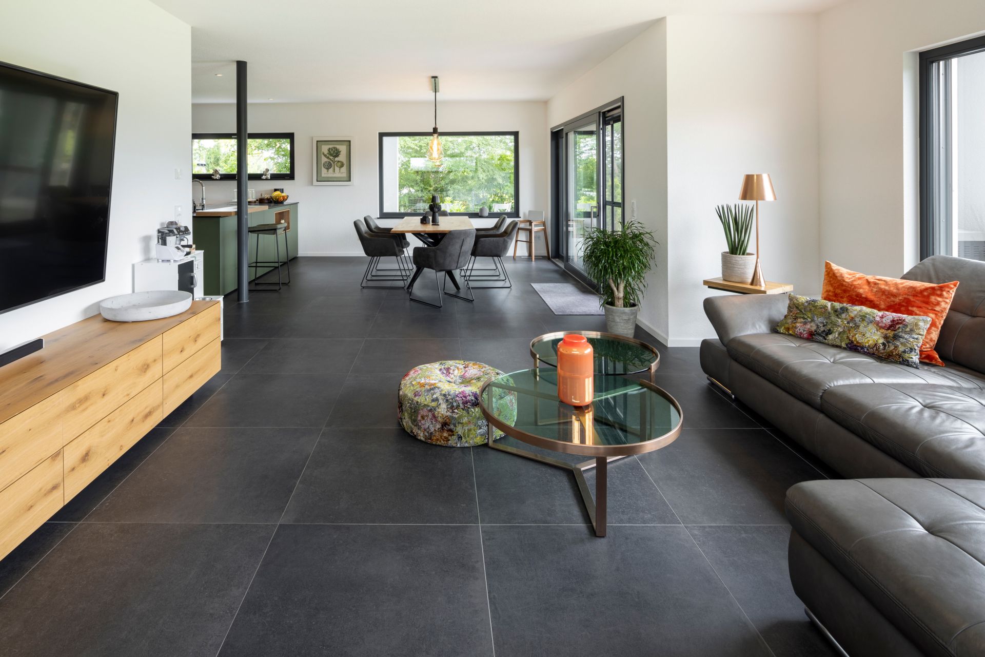 Offener Wohn- und Essbereich in einem modernen FingerHaus-Haus mit grauen Bodenfliesen, Ledersofa, Glas-Couchtischen, stilvollen Dekorationen und großen Fenstern mit Blick ins Grüne.