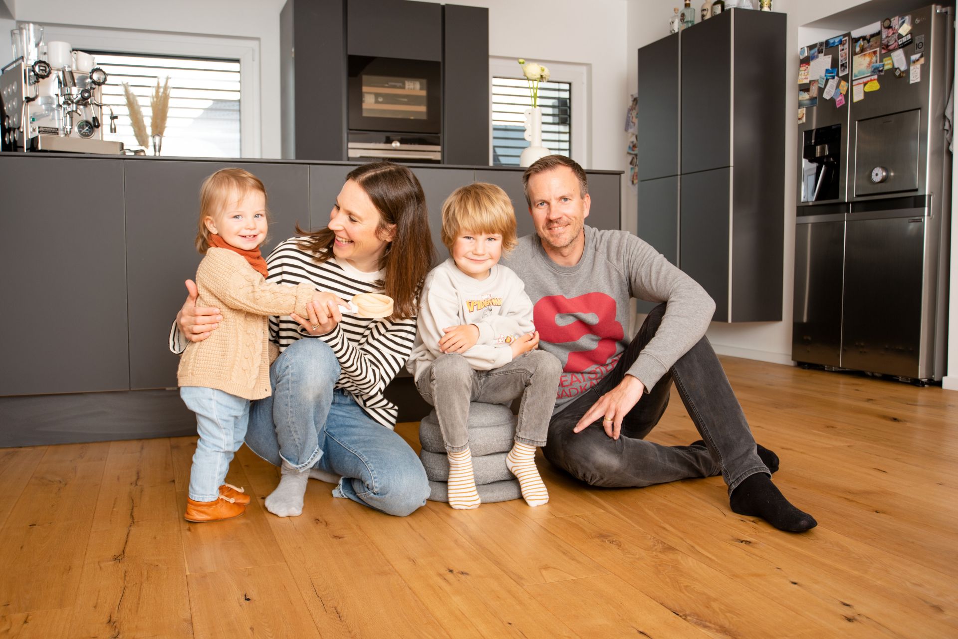 Eine Familie mit zwei kleinen Kindern sitzt lachend auf dem Holzboden ihrer modernen Küche in einem FingerHaus-Haus. Im Hintergrund sind dunkle Küchenschränke und Geräte zu sehen.