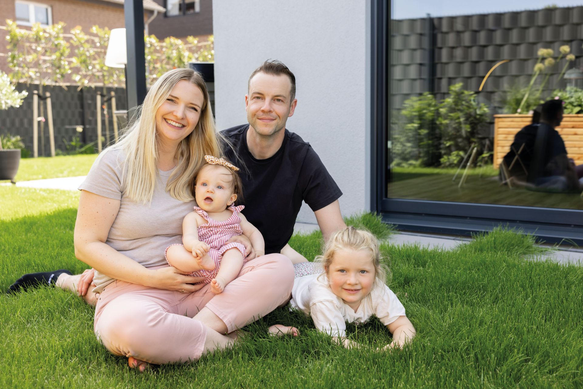 Familie mit zwei Kindern sitzt gemeinsam im Garten auf dem Rasen vor ihrem Haus