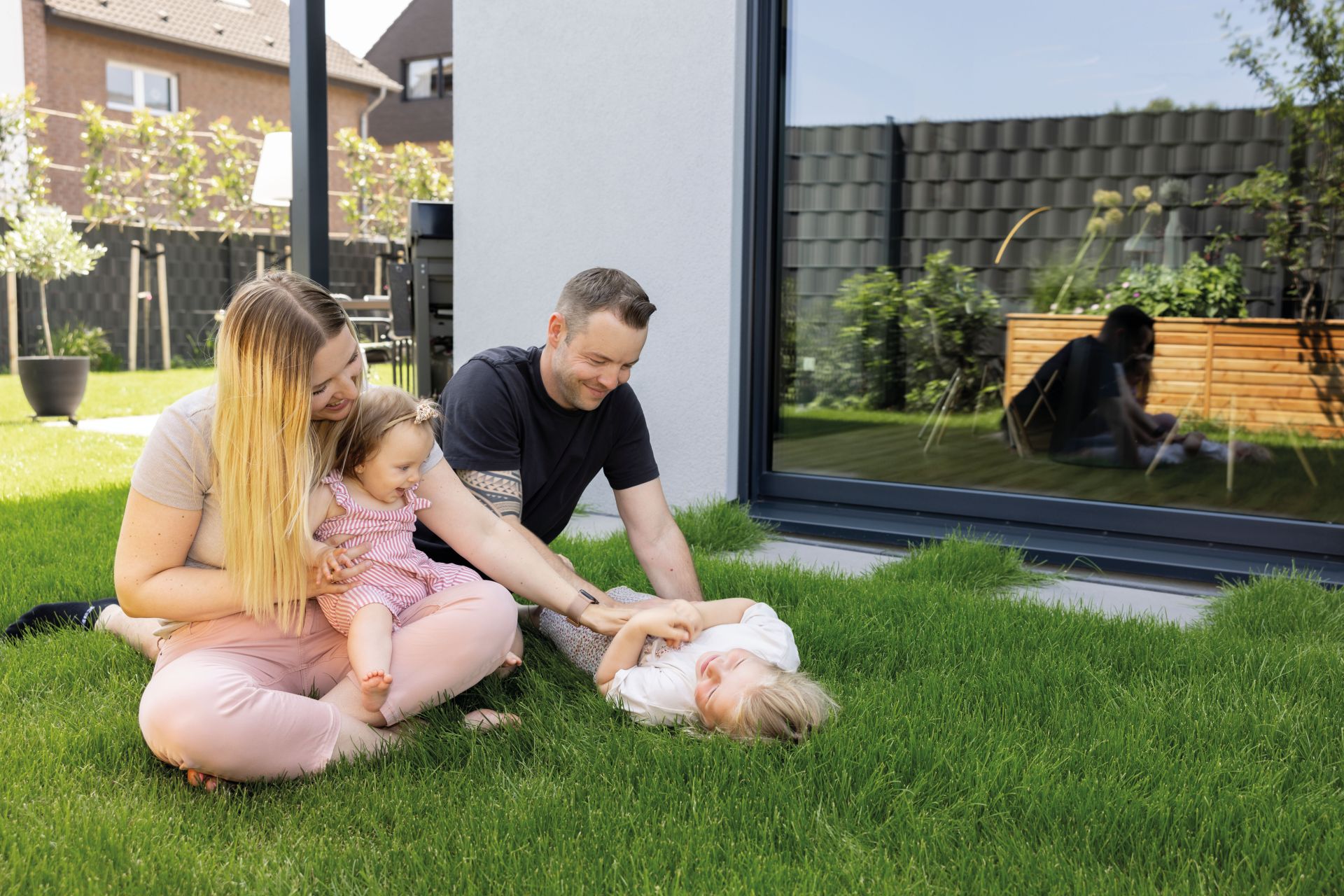 Familie mit zwei Kindern sitzt und spielt auf dem Rasen im Garten vor einer großen Fensterfront