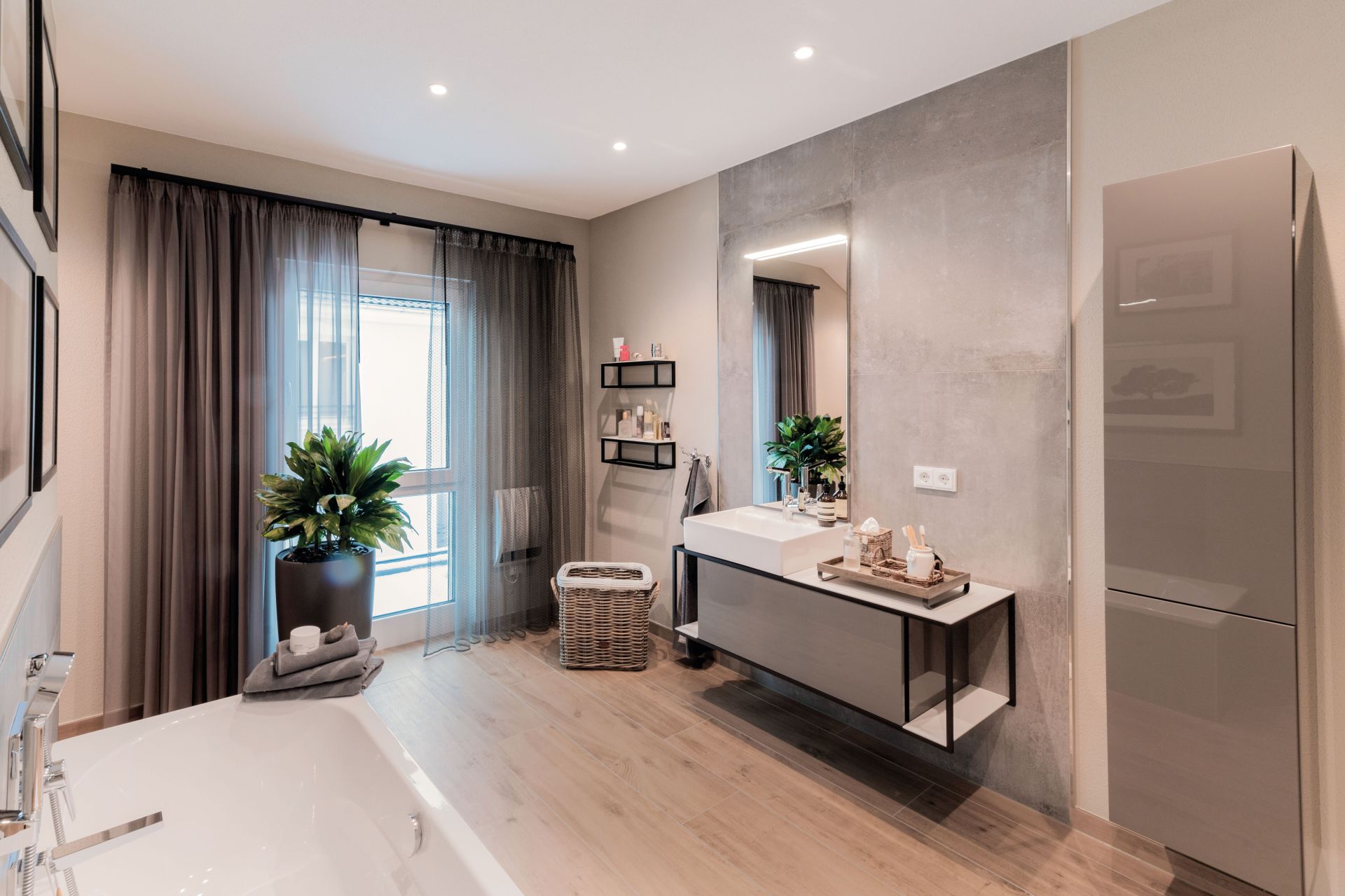 Modern eingerichtetes Badezimmer mit freistehender Badewanne, großem Spiegel über einem wandhängenden Waschbecken, Pflanzen und einem Wäschekorb vor bodentiefen Fenstern mit Vorhängen