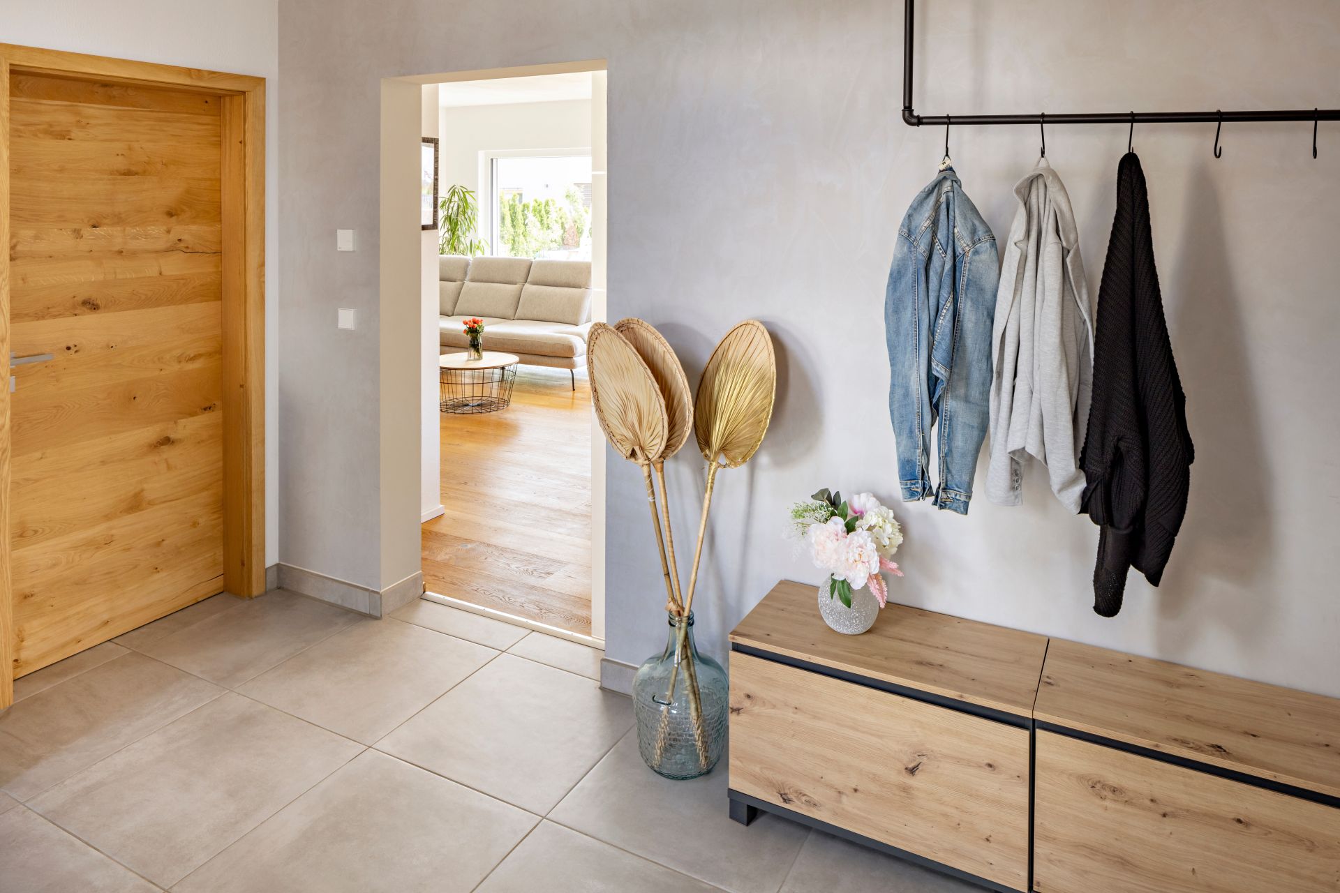 Eingangsbereich mit Holzschrank, schwarzer Kleiderstange mit Jacken, grauem Fliesenboden und einer Holztür; durch den Durchgang ist das Wohnzimmer sichtbar.