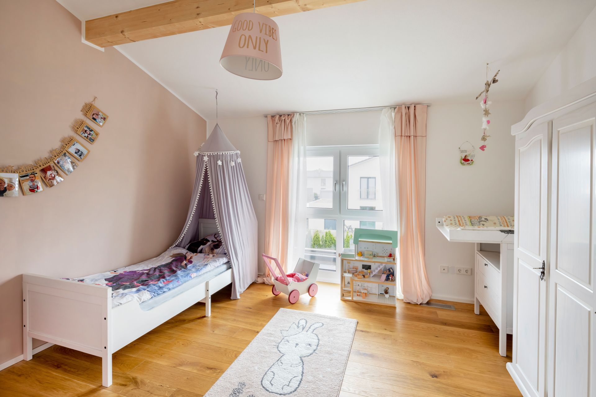 Kinderzimmer mit weißem Einzelbett unter einem Stoffhimmel, Spielküche, Puppenwagen, Wickelkommode, Kleiderschrank und großem Fenster.