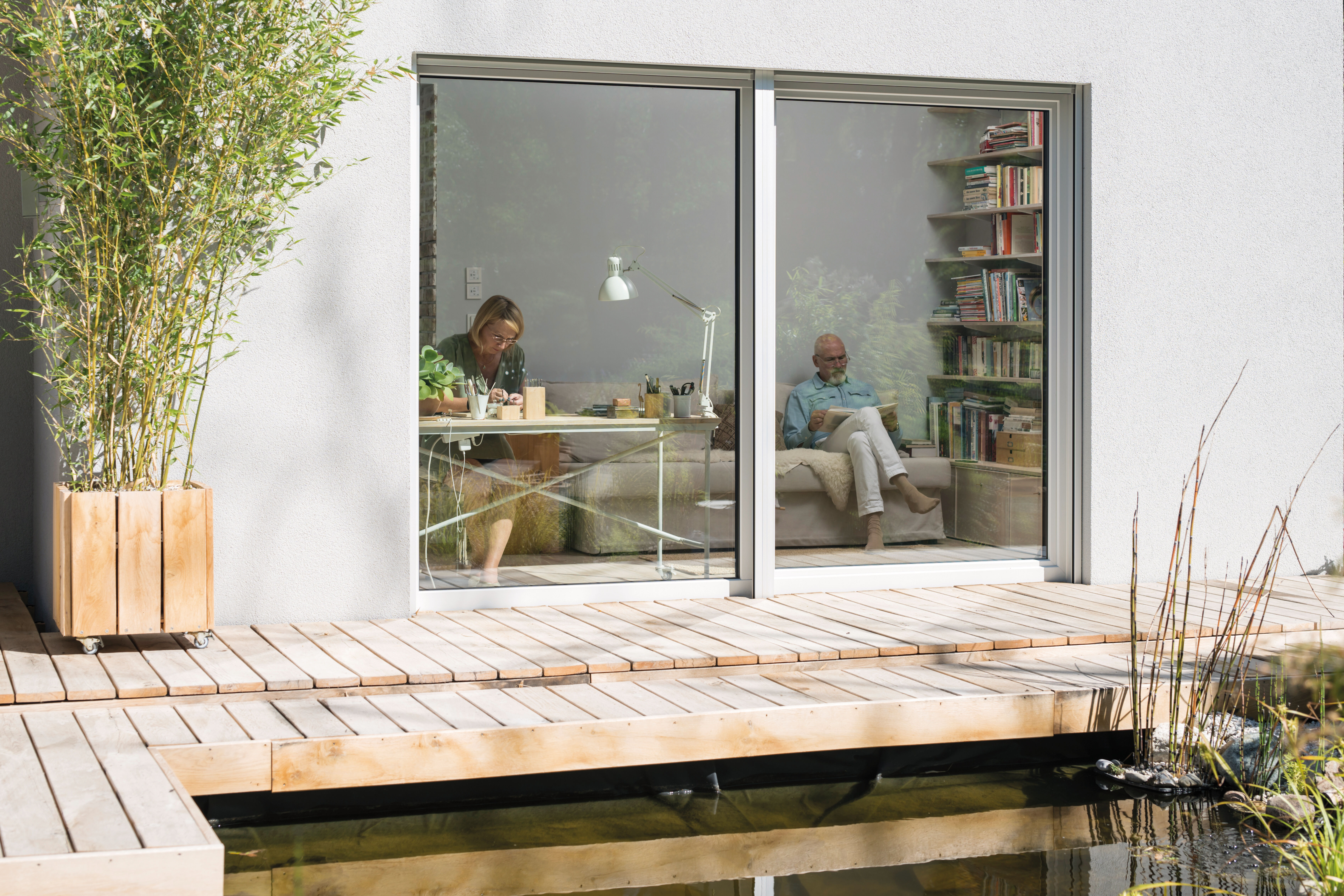 Bungalow mit großer Fensterfront und Holzterrasse am Wasser, innen ein Paar beim Arbeiten und Lesen