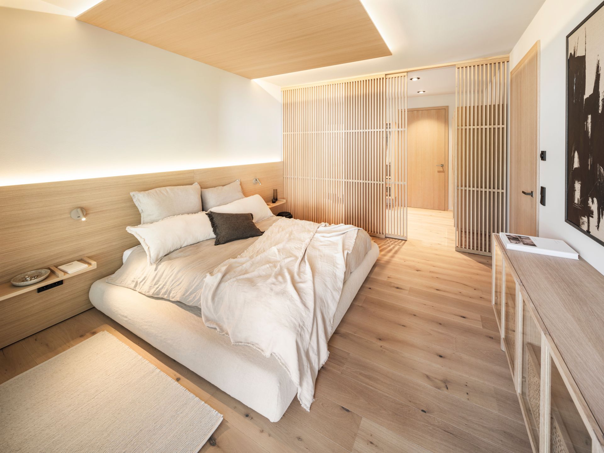 Schlafzimmer im FingerHaus Musterhaus NIVO in Frankenberg mit Doppelbett, Holzfußboden, indirekter Beleuchtung und Raumteiler aus Holzlamellen.