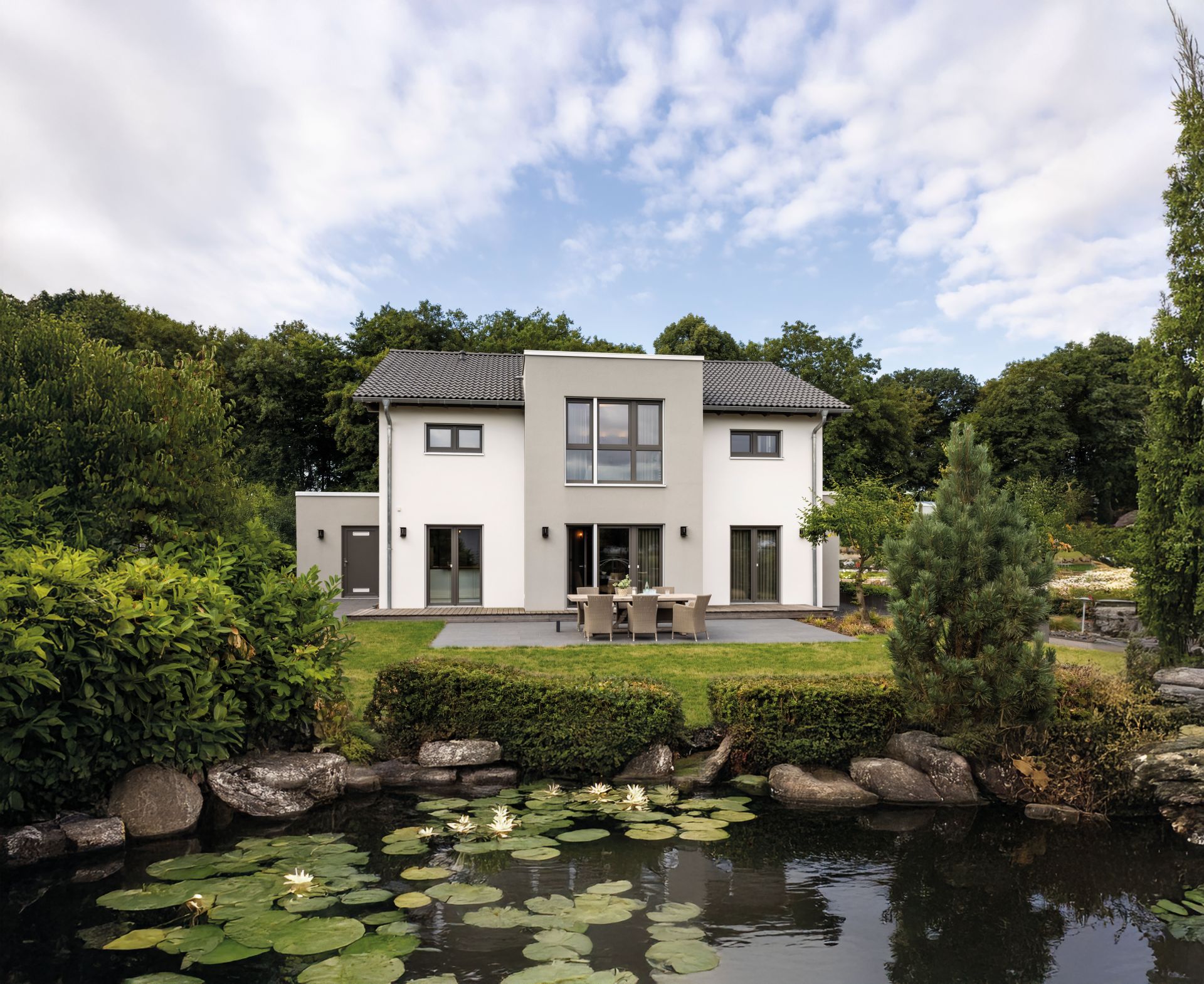 Zweigeschossiges Wohnhaus mit heller Putzfassade, symmetrischem Satteldach, mittigem Fassadenvorsprung mit großen Fenstern, Terrasse mit Tisch und Stühlen sowie Rasenfläche im Garten.