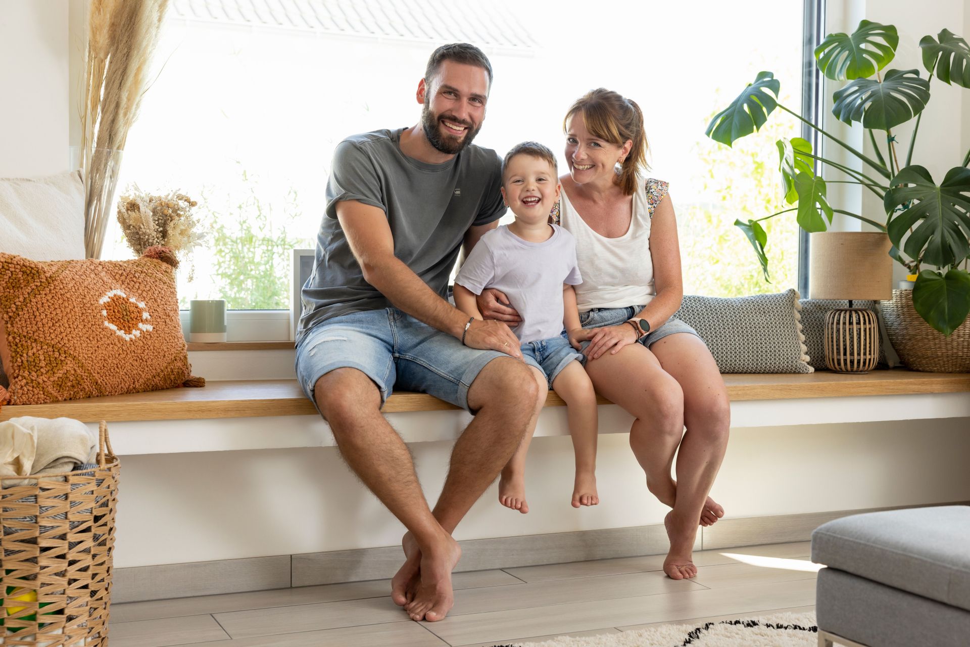 Familie mit zwei Erwachsenen und einem Kind sitzt auf einer Fensterbank im Wohnbereich und lächelt in die Kamera