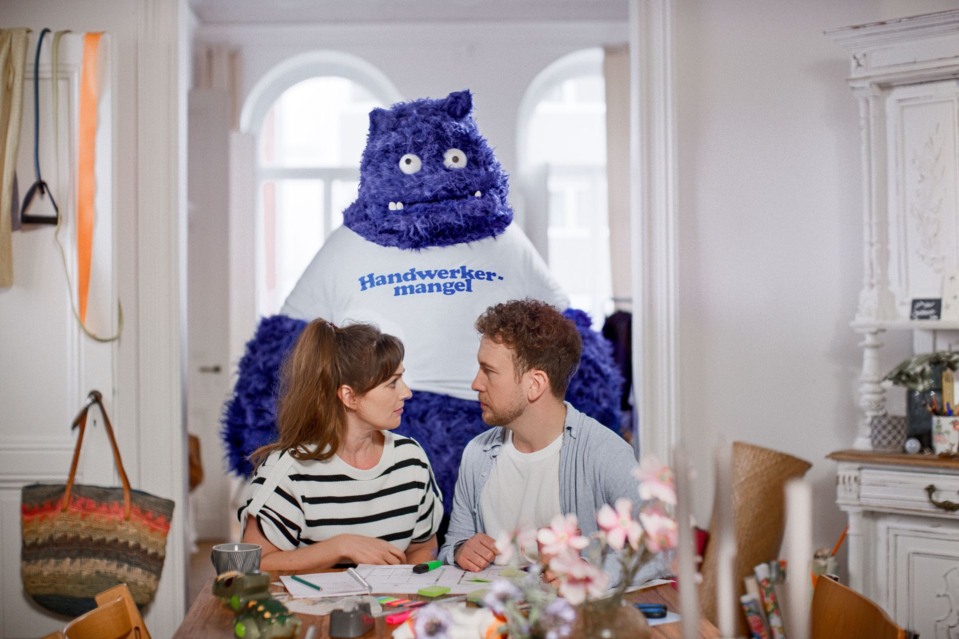 Ein Paar sitzt am Tisch und bespricht Baupläne, während hinter ihnen ein großes blaues, plüschartiges Wesen mit der Aufschrift „Handwerkermangel“ auf dem T-Shirt steht – Symbolfigur aus der Kampagne „Das Unerwartete“.
