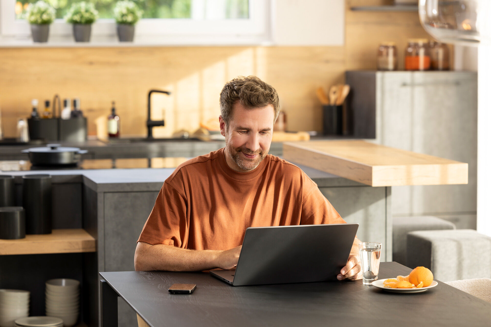 Mann sitzt an einem Tisch mit Laptop in einer modernen Küche mit Kücheninsel, Holzarbeitsplatte, Spüle, Hängelampe, Glas Wasser, Teller mit Orangen, Smartphone und Barhockern im Hintergrund.