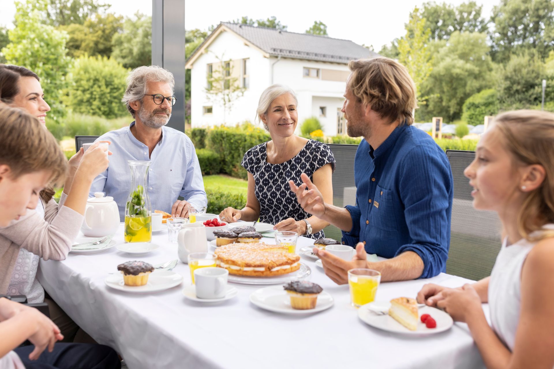 Mehrere Personen sitzen an einem gedeckten Tisch auf einer Terrasse und genießen gemeinsam Kaffee und Kuchen vor einem Wohnhaus im Grünen.