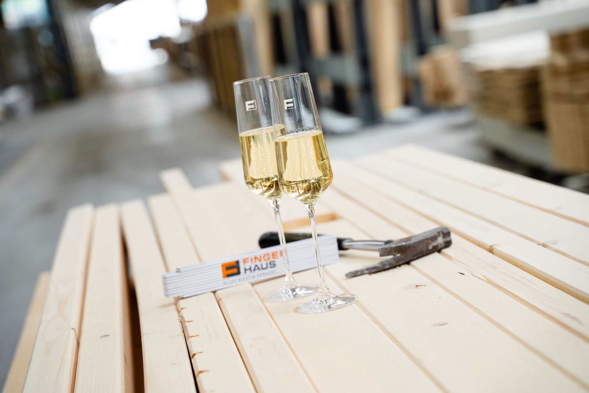 Zwei gefüllte Sektgläser stehen auf hellen Holzlatten in einer Werkhalle, daneben ein Zollstock mit FingerHaus-Logo und ein Hammer.