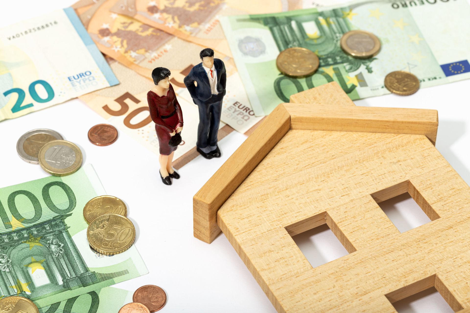 Miniaturfiguren eines Paares stehen auf Euro-Banknoten und Münzen neben einem Holzhausmodell – Symbolbild für Immobilienkauf und Finanzierung.