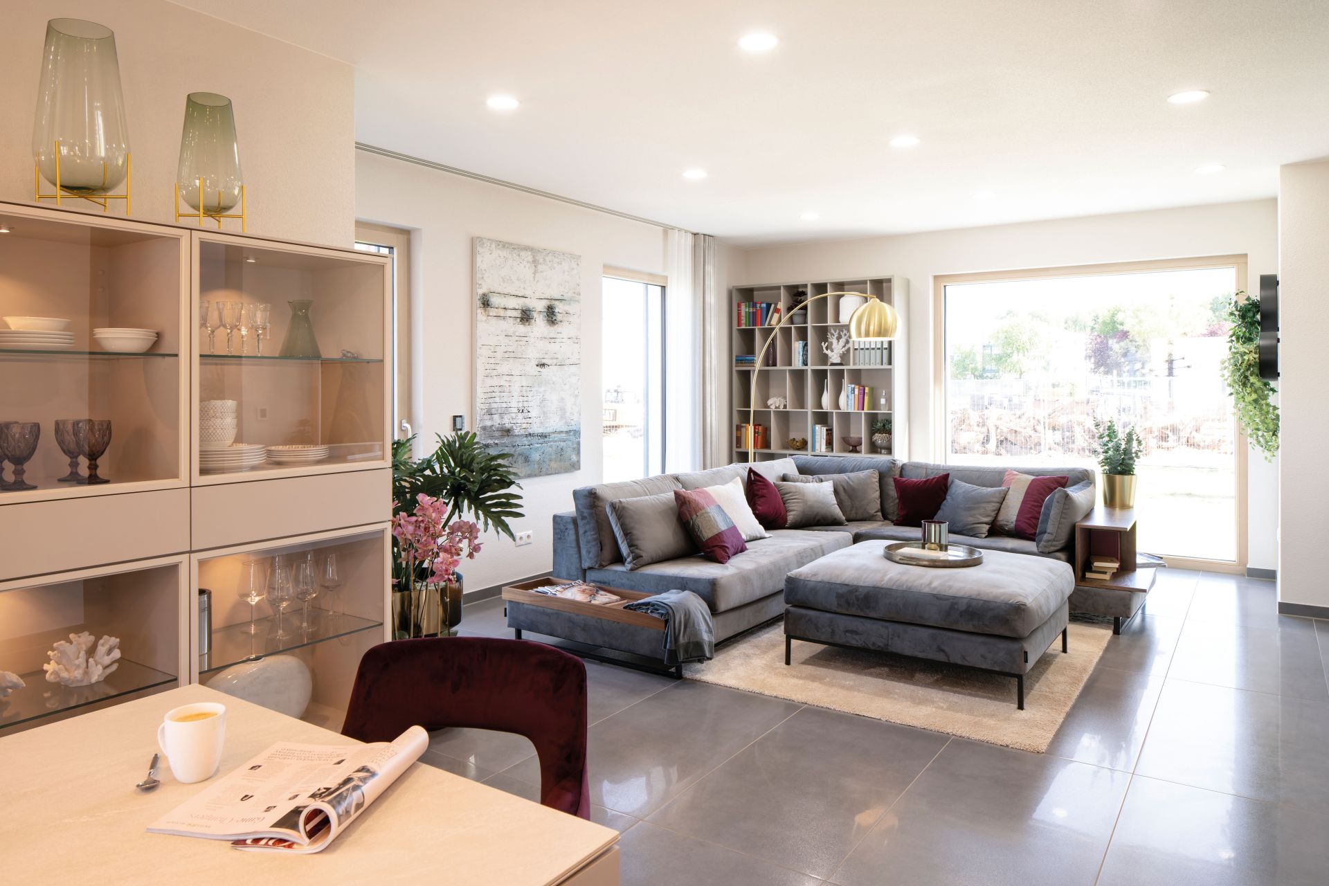 Wohnbereich im Musterhaus MEDLEY 300 B in Frankenberg mit großem grauen Ecksofa, Glastürschrank, moderner Deko und Panoramafenster.