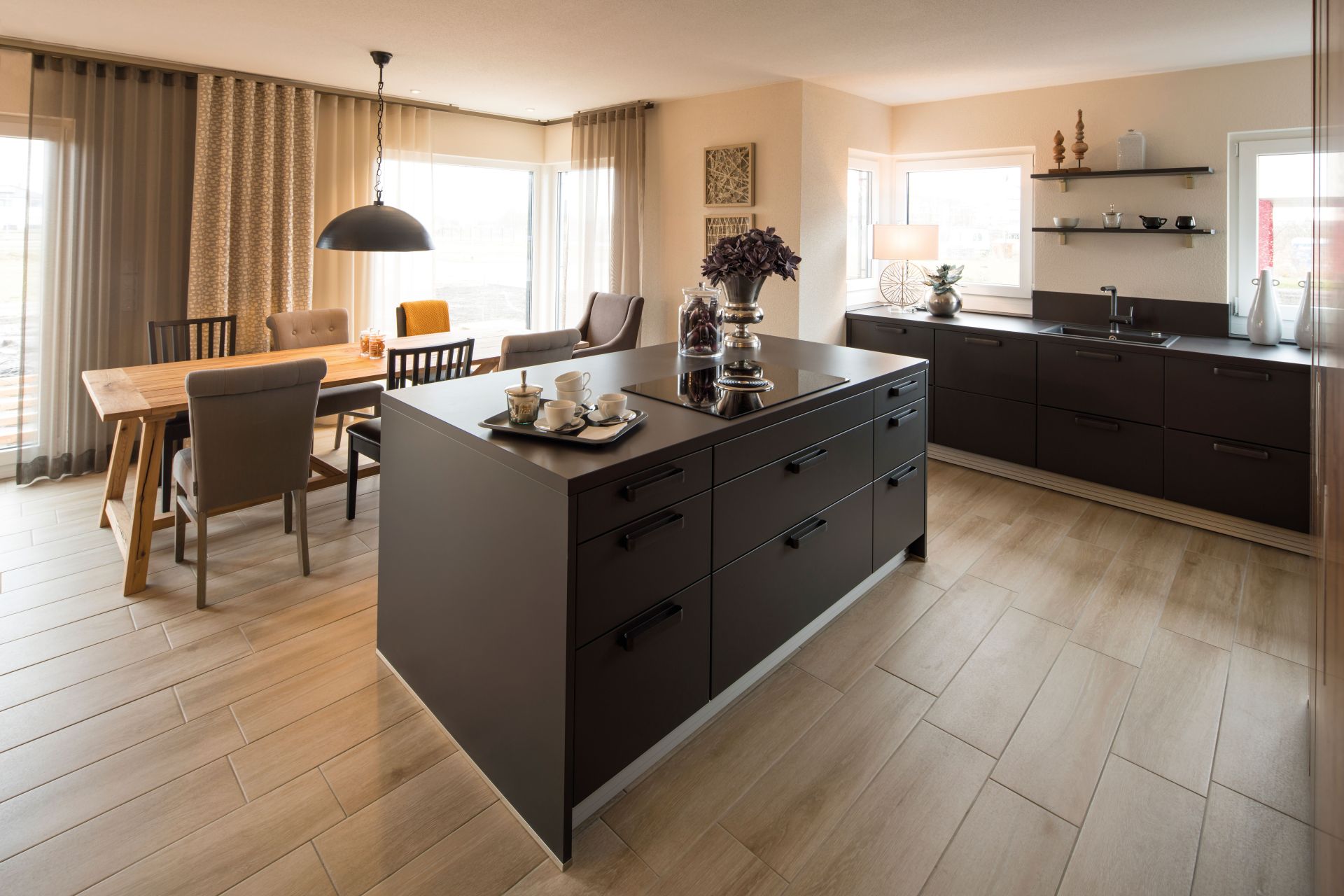 Essbereich und Küche im FingerHaus MEDLEY 300 B Musterhaus Berlin/Werder mit dunkler Kücheninsel, Esstisch aus Holz und großen Fenstern.