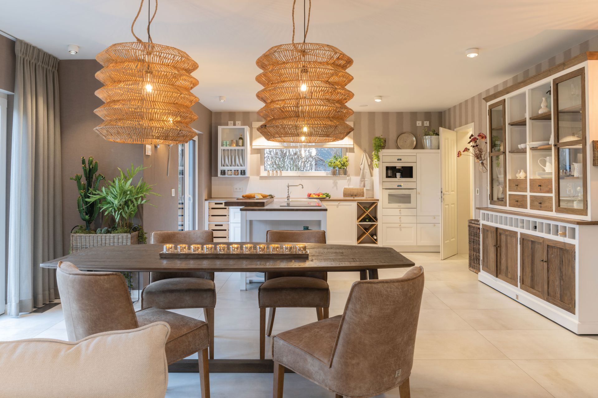 Gemütlicher Essbereich im Musterhaus SENTO von FingerHaus in Bad Vilbel mit großem Holztisch, Lederstühlen, markanten Korbleuchten und angrenzender Landhausküche mit Vitrine.