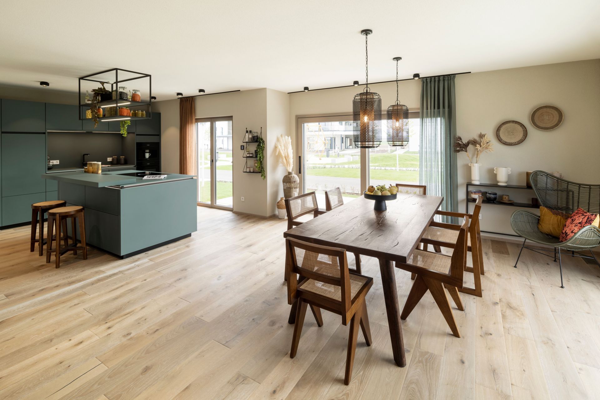 Offene Küche mit Essbereich im FingerHaus ARTIS 400 Musterhaus Schwarzwald mit moderner Kücheninsel in Grün, großem Holztisch und stilvollen Leuchten.