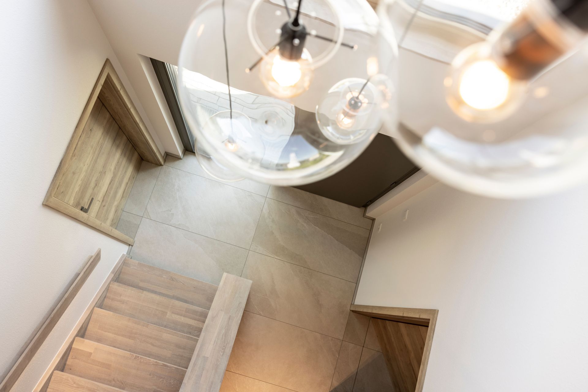 Blick von oben auf eine moderne Holztreppe mit großformatigen Fliesen im Eingangsbereich des Musterhaus ARTIS in Nürnberg. Große Fenster und kugelförmige Pendelleuchten sorgen für helles, natürliches Licht.