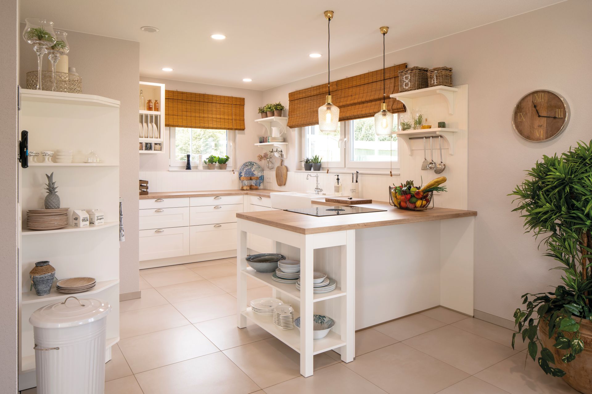Helle Landhausküche mit weißer Kochinsel, offenen Regalen und Holzarbeitsplatte im Musterhaus JUNO Frankenberg. Große Fenster mit Raffrollos, Hängelampen und Pflanzen verleihen dem Raum eine freundliche Atmosphäre.