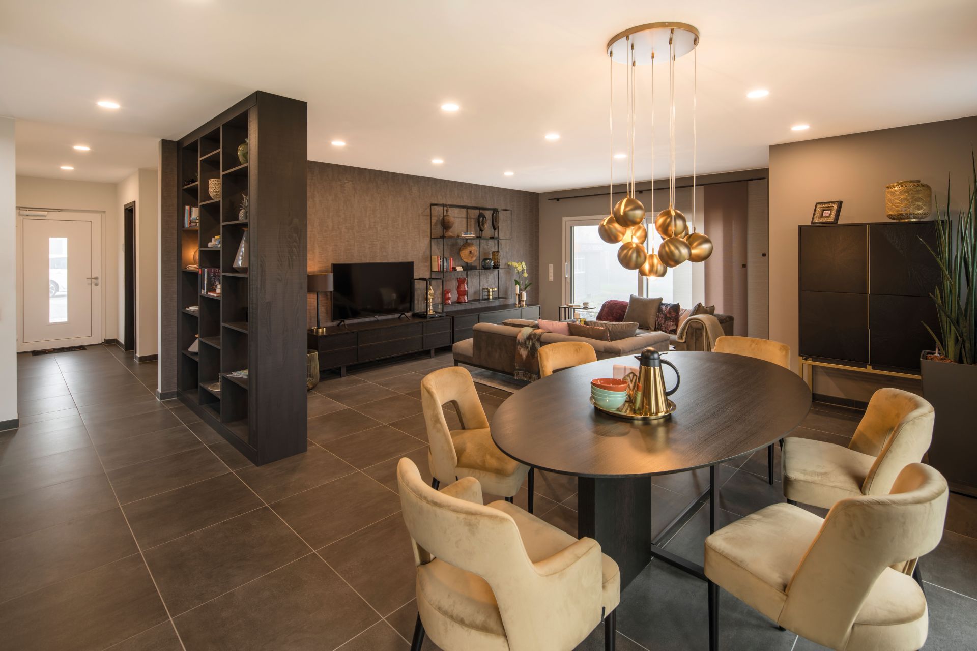 Innenansicht des Musterhauses MEDLEY 300 B in Kassel mit Essbereich, rundem Tisch, samtbezogenen Stühlen und modernem Wohnzimmer im Hintergrund.