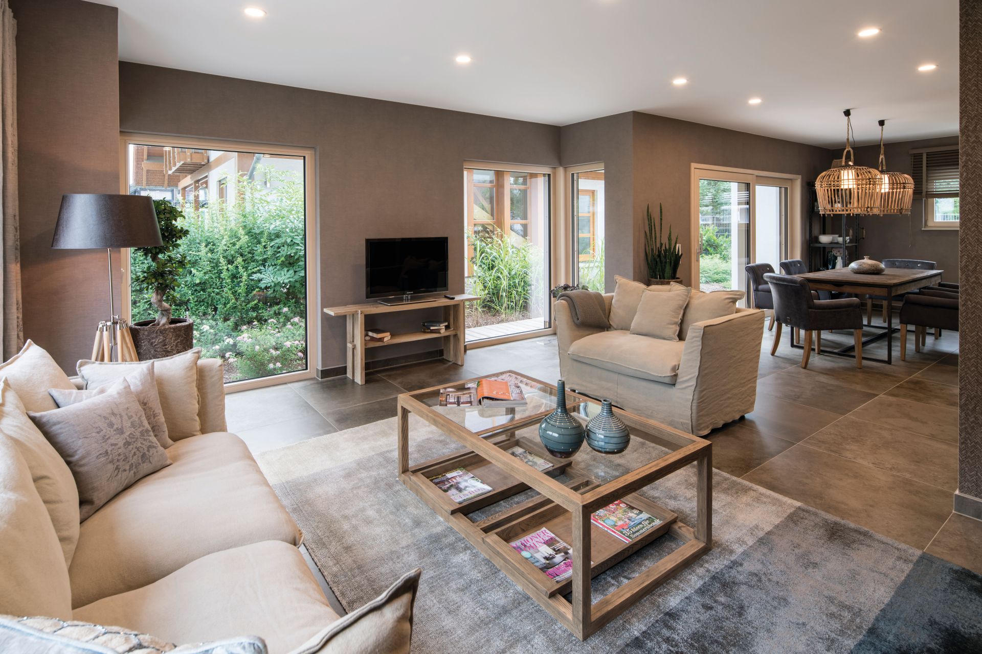 Wohnzimmer im Musterhaus MEDLEY in Fellbach mit großen Fenstern, gemütlichen Sofas, Holztisch und angrenzendem Essbereich.