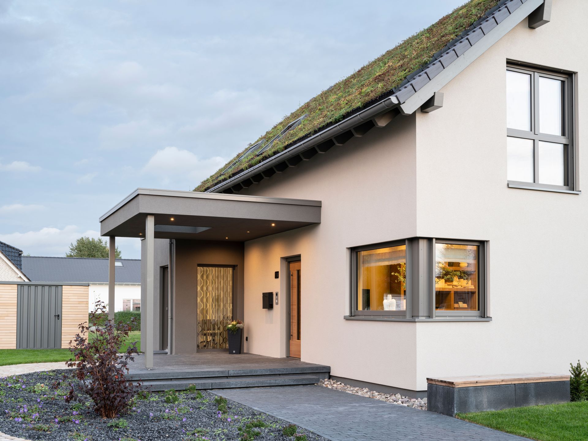 Moderner Eingangsbereich des Musterhauses JUNO 300 in Mülheim-Kärlich mit überdachter Terrasse, Holzdetails und begrüntem Dach.