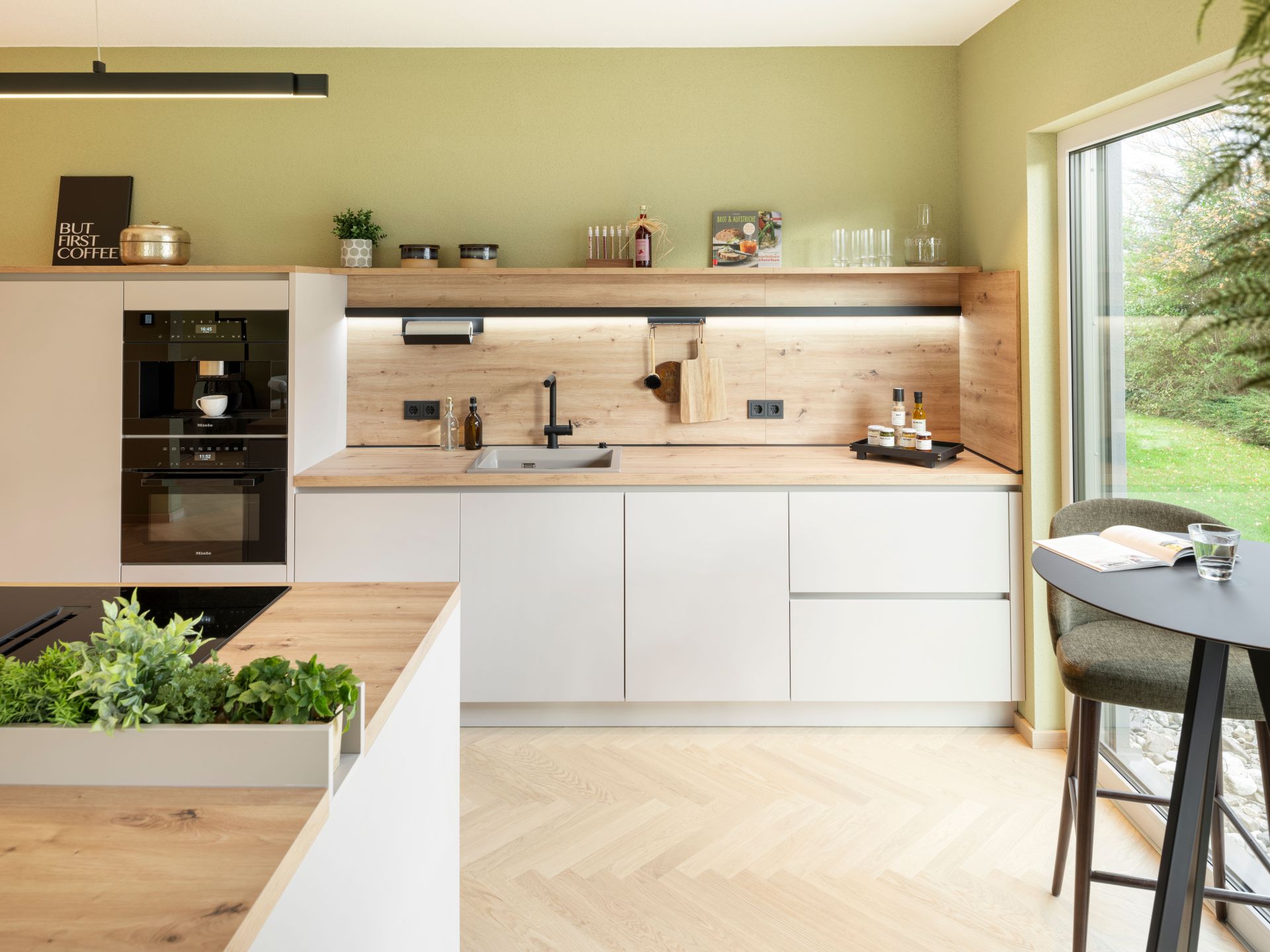 Moderne Küche im Musterhaus JUNO 300 in Mülheim-Kärlich mit weißen Fronten, Holzarbeitsplatten, integriertem Kräuterbeet und Essplatz am Fenster.