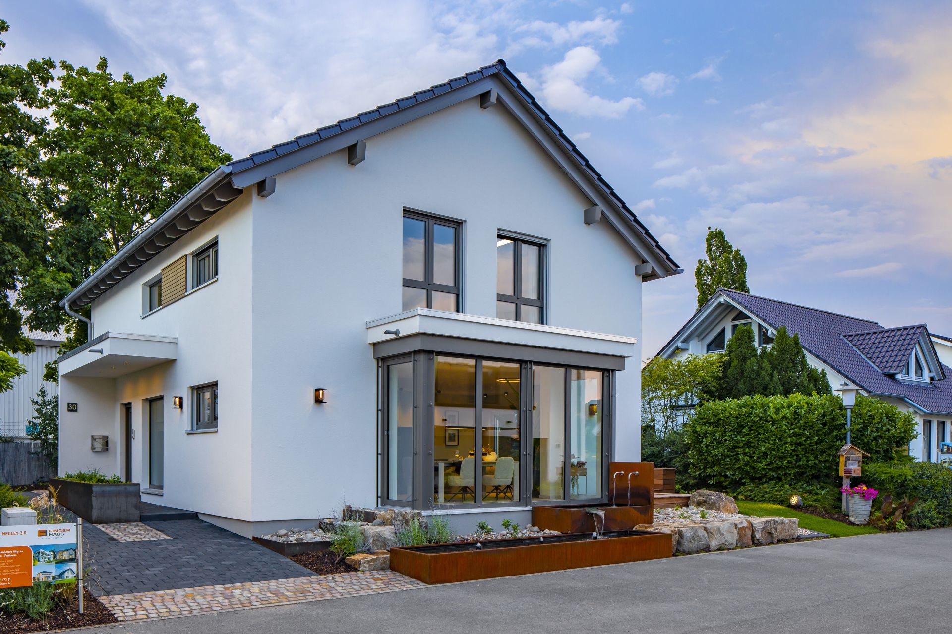 Abendstimmung am Musterhaus MEDLEY in Mannheim mit weißer Fassade, großen Fensterfronten und stilvoller Außenanlage.