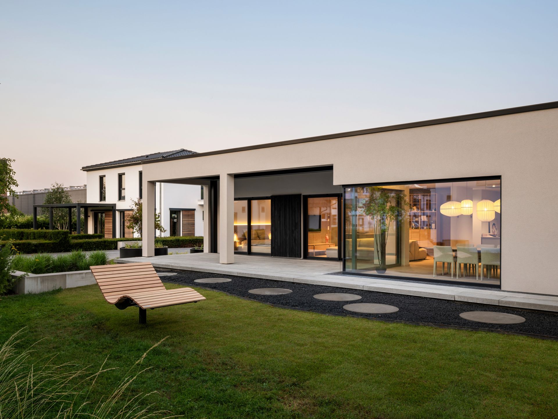 Musterhaus NIVO in Frankenberg mit moderner Bungalowarchitektur, überdachter Terrasse, großzügigen Fensterfronten und beleuchtetem Wohn- und Essbereich in der Abenddämmerung.