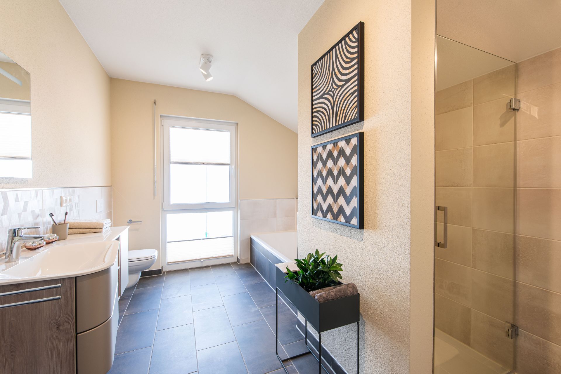 Modernes Badezimmer im FingerHaus Musterhaus TALO in Babenhausen mit Dusche, Badewanne, Waschtisch und dekorativen Wandbildern. Helle Fliesen kombiniert mit dunklem Bodenbelag und stilvollen Einrichtungselementen.