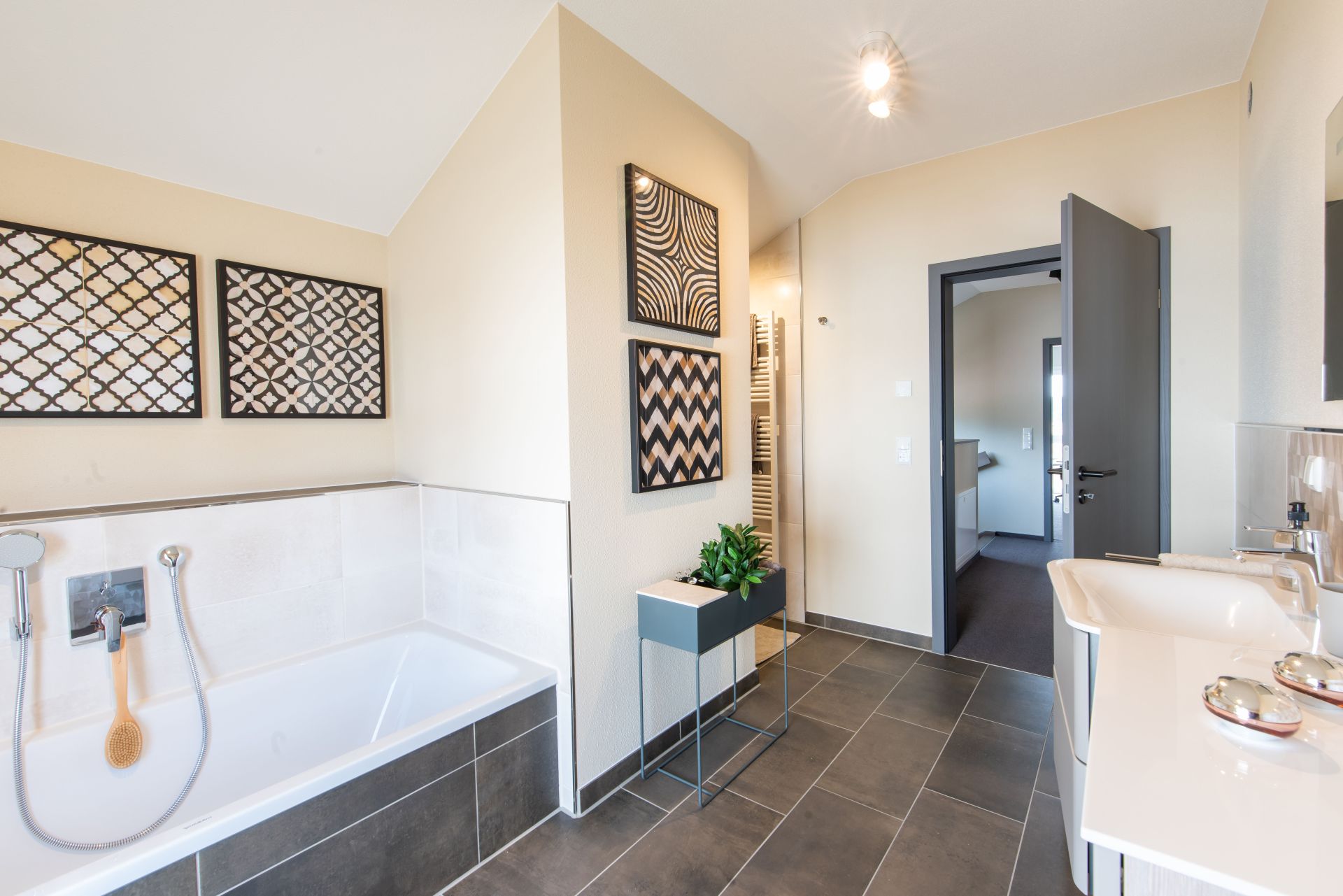 Modernes Badezimmer im FingerHaus Musterhaus TALO in Babenhausen mit Badewanne, Doppelwaschbecken und dekorativen Wandbildern. Helle Wandgestaltung kombiniert mit dunklem Boden und stilvollen Einrichtungselementen.