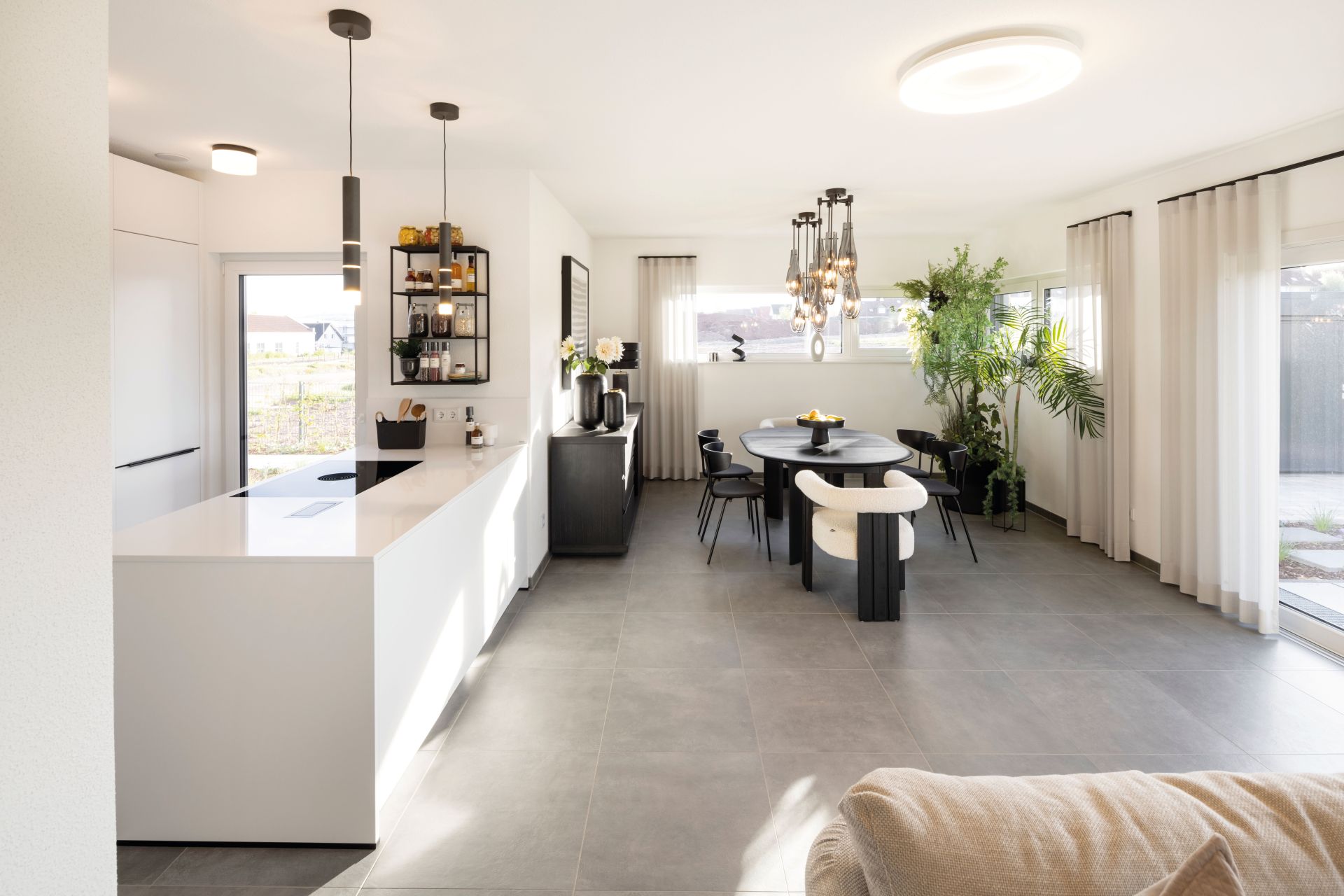 Das Musterhaus TALO in Fulda zeigt eine helle Wohnküche mit großzügigem Essbereich und stilvollem Design. Offener Küchen- und Essbereich mit Kücheninsel, Esstisch, Stühlen, Pendelleuchten, Pflanzen, Fensterfront mit Vorhängen