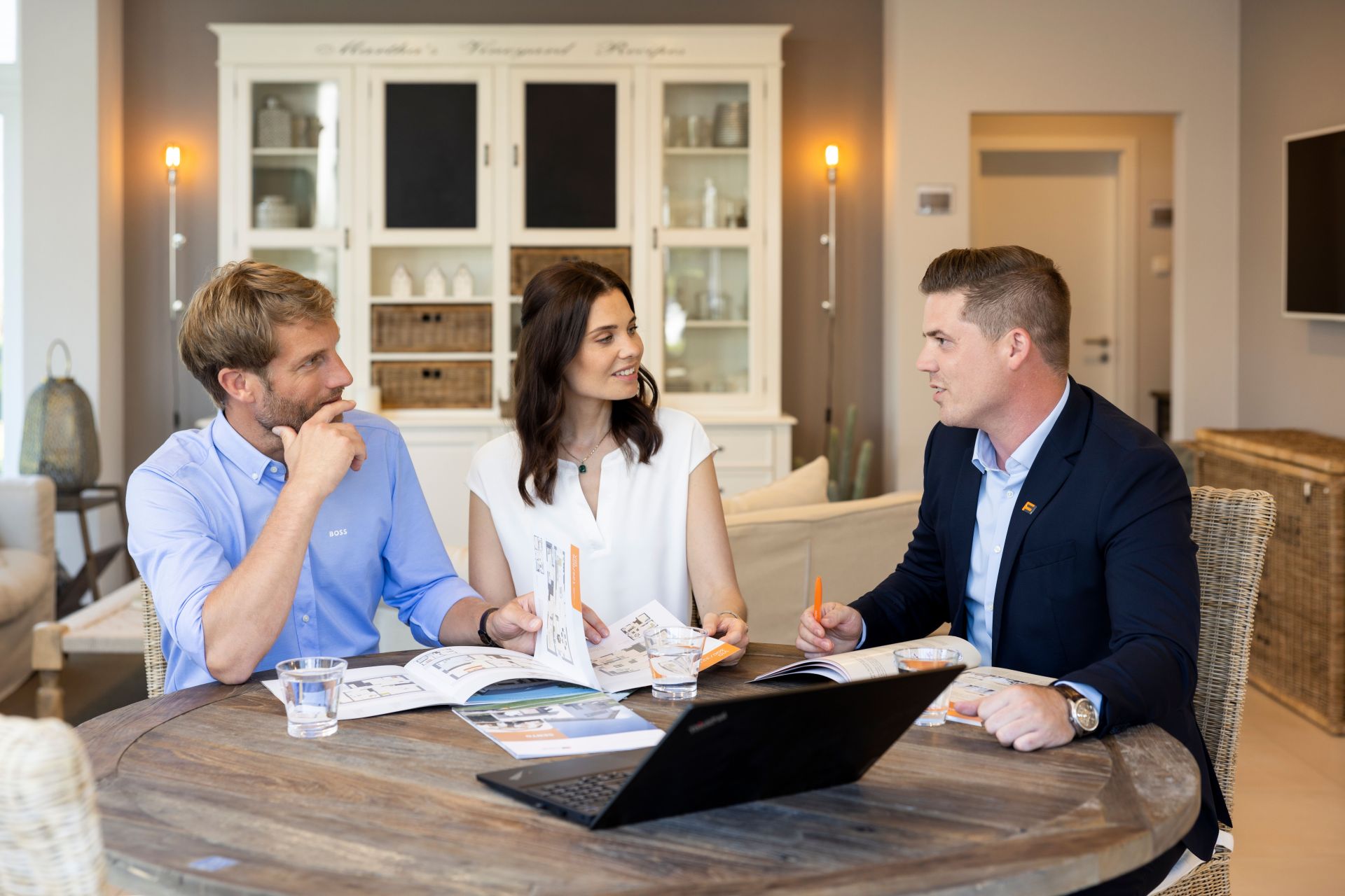 Beratungssituation mit einem FingerHaus-Fachberater: Ein Paar sitzt gemeinsam mit dem Berater an einem Tisch, betrachtet Unterlagen und Kataloge und bespricht die Planung eines Finger-Hauses in wohnlicher Atmosphäre.