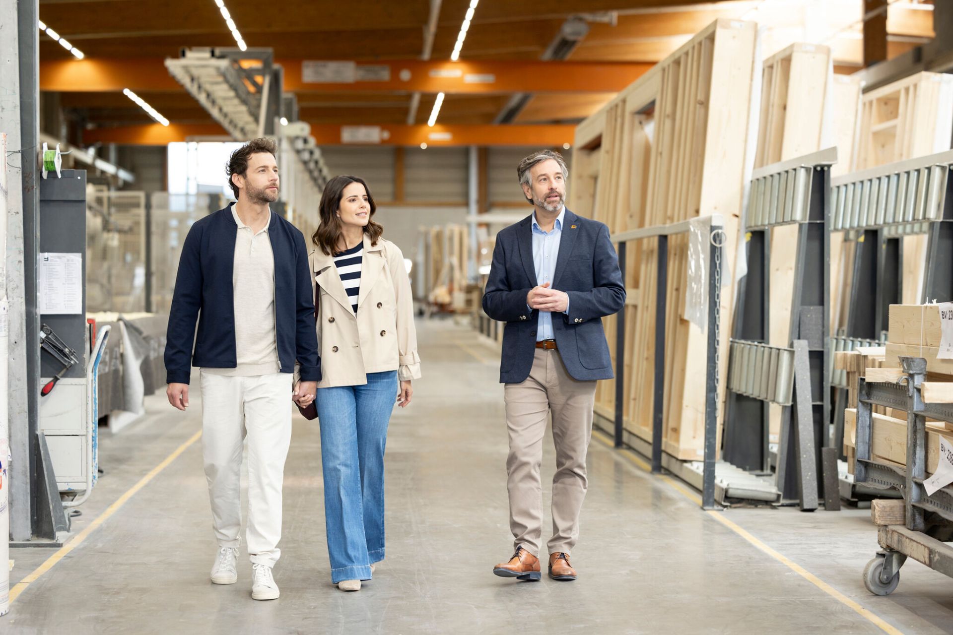 Zwei erwachsene Personen und ein FingerHaus Fachberater gehen gemeinsam durch eine Produktionshalle mit vorgefertigten Holzbauelementen und Wandmodulen.  